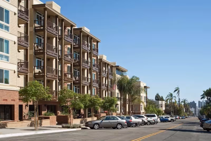 Street view of a multi-story residential building with balconies, trees lining the sidewalk, and parked cars along the street under a clear blue sky.