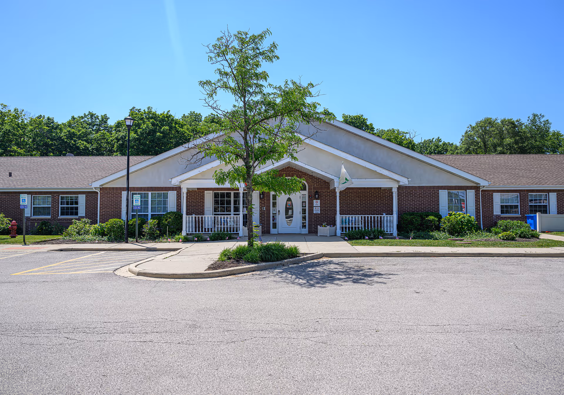 Front exterior view of a single-story brick building with a white entrance porch, a small tree in front, and a clear blue sky. The building has multiple windows and a parking area with handicap parking spaces.