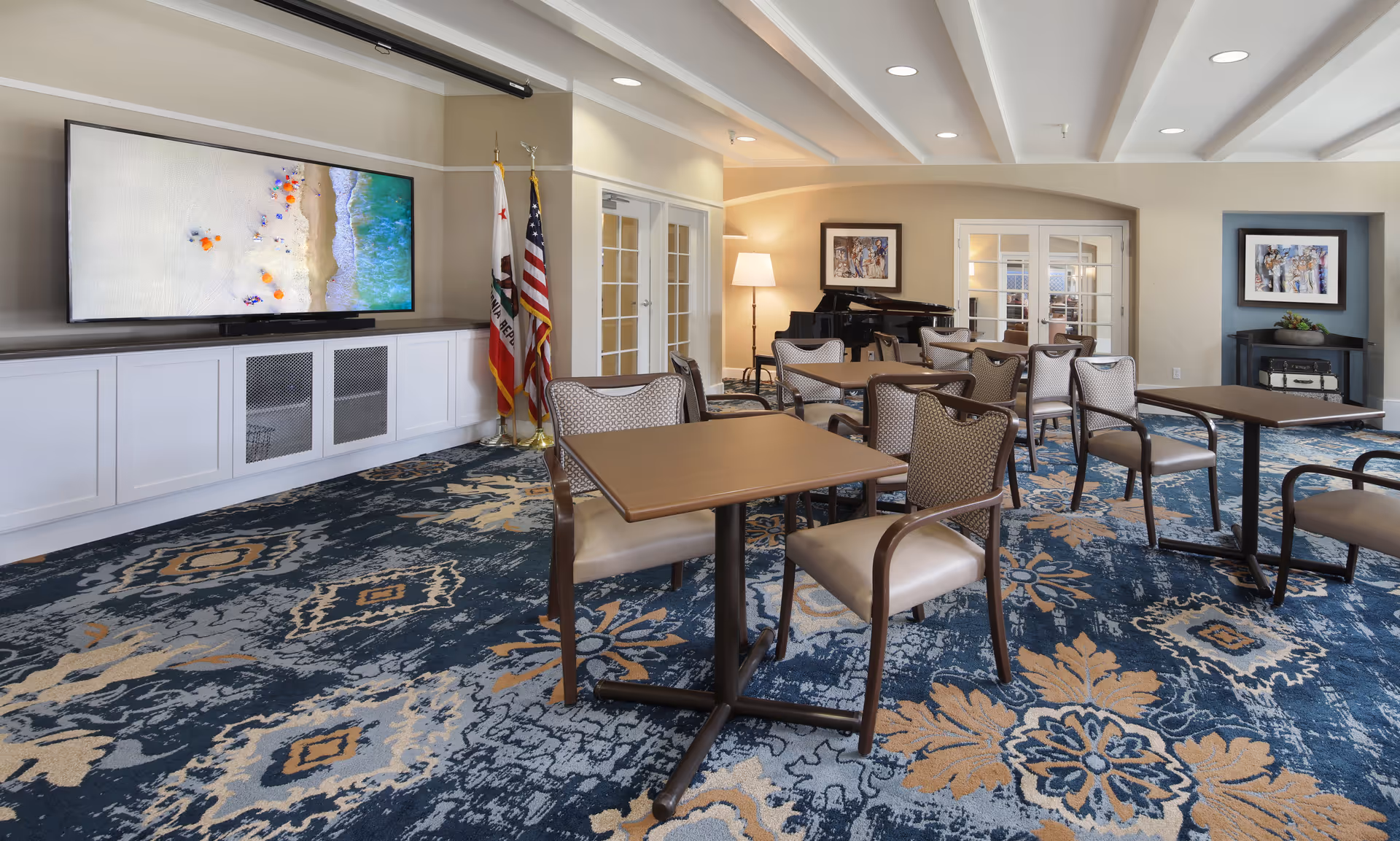 A spacious room with multiple tables and chairs arranged on a patterned blue and beige carpet. A large flat-screen TV mounted on the wall displays a beach scene. Two flags stand next to the TV. In the background, there is a black grand piano, a floor lamp, framed artwork on the walls, and glass double doors.