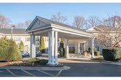 Exterior view of Sterling Care Bethesda showing the entrance with a covered driveway supported by white columns, surrounded by bushes and trees under a clear sky.