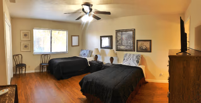 A senior care facility bedroom with two single beds covered in black bedspreads, each with a decorative pillow. Between the beds is a nightstand with two matching lamps. The room has wooden flooring, a ceiling fan with lights, a window with blinds, two chairs near the window, and framed artwork on the walls. A wooden dresser with a TV on top is visible on the right side of the room.