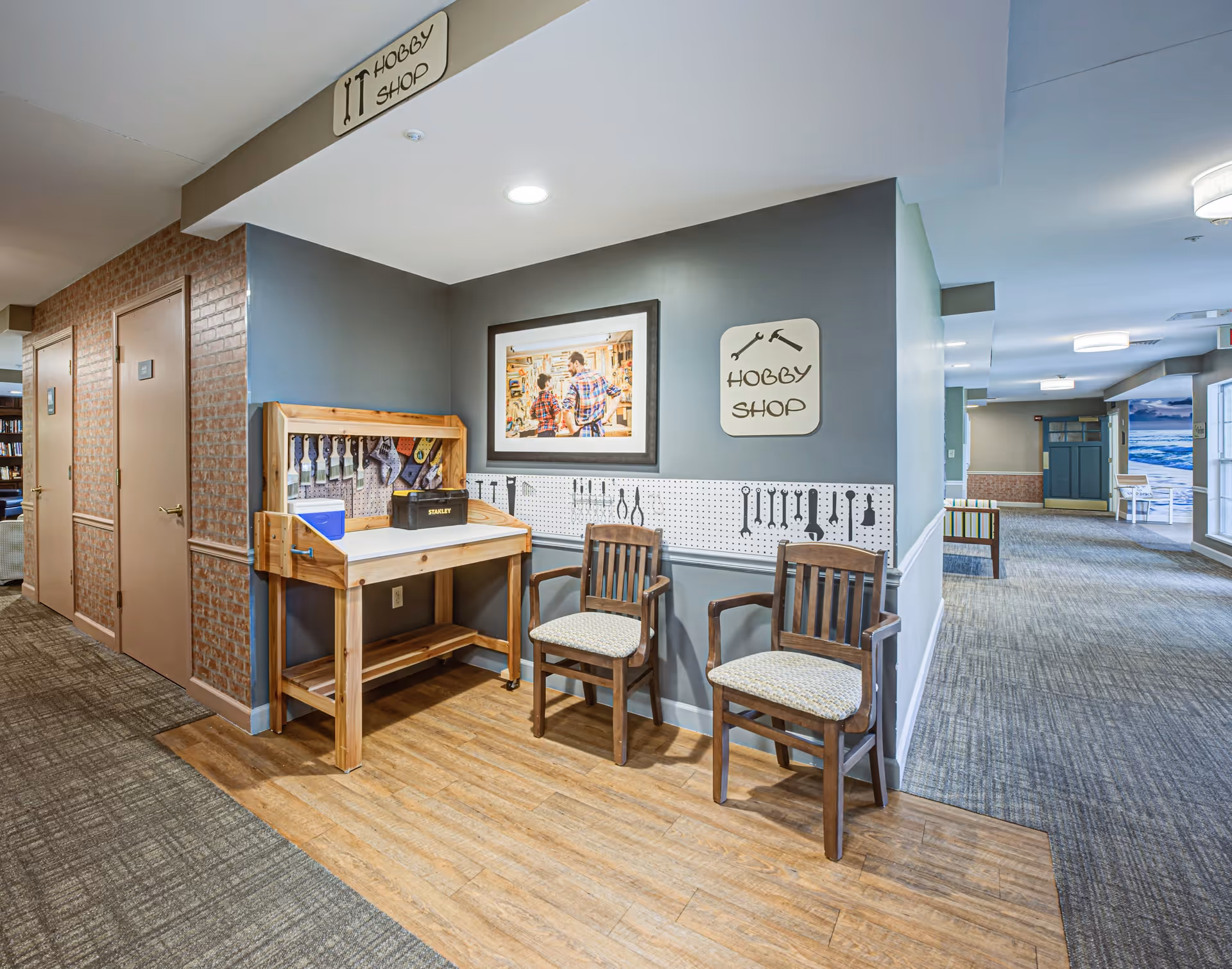 Interior hallway area of Arden Courts - ProMedica Memory Care Community featuring a hobby shop corner with a wooden workbench, tools hanging on a pegboard, two wooden chairs with cushions, and a framed picture on the wall. The hallway has carpeted flooring and leads to other rooms and seating areas.
