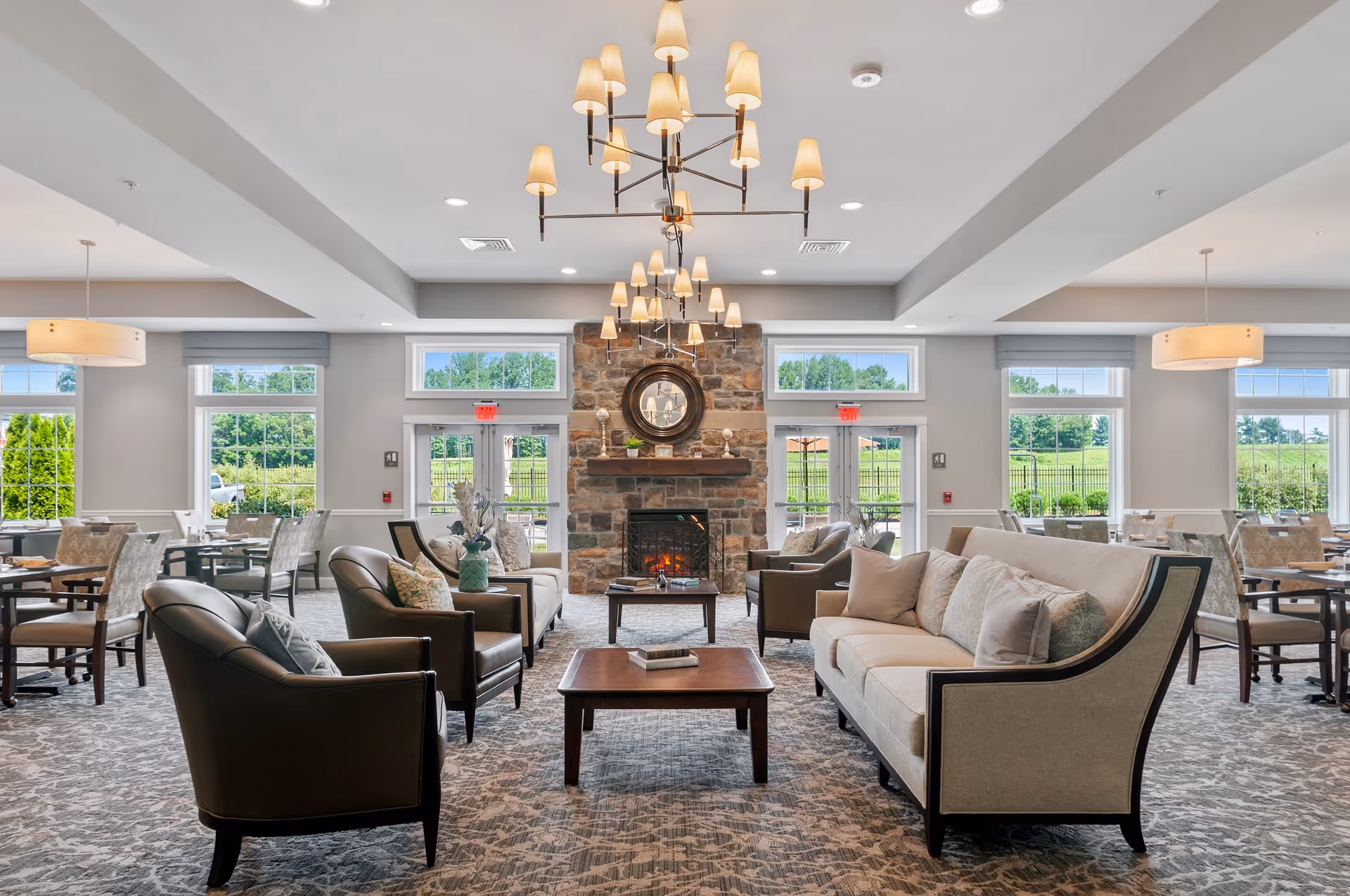 A spacious and well-lit senior living common area featuring comfortable beige sofas and dark leather armchairs arranged around a wooden coffee table. A stone fireplace with a round mirror above it is centered on the far wall. Large windows and glass doors let in natural light and offer views of green outdoor spaces. Multiple chandeliers and pendant lights hang from the ceiling, illuminating the room. Dining tables and chairs are visible in the background.