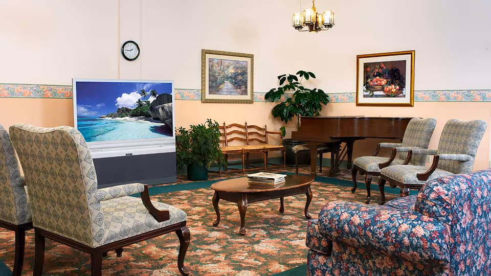 A cozy living room area with patterned armchairs and a floral sofa arranged around a wooden coffee table with books. A large television displays a beach scene, and a grand piano is positioned against the wall. The room features framed artwork, a clock, potted plants, and a floral carpet.
