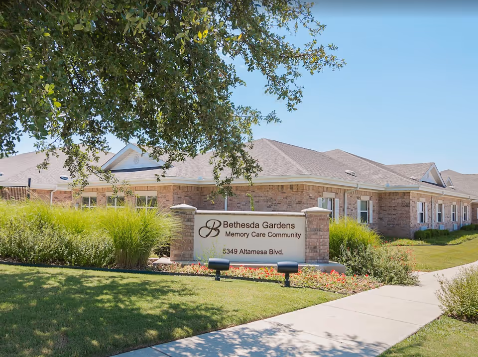 Exterior view of Bethesda Gardens Memory Care Community building with a brick facade, surrounded by green grass, bushes, and a tree. A sign in front displays the facility name and address: 5349 Altamesa Blvd.