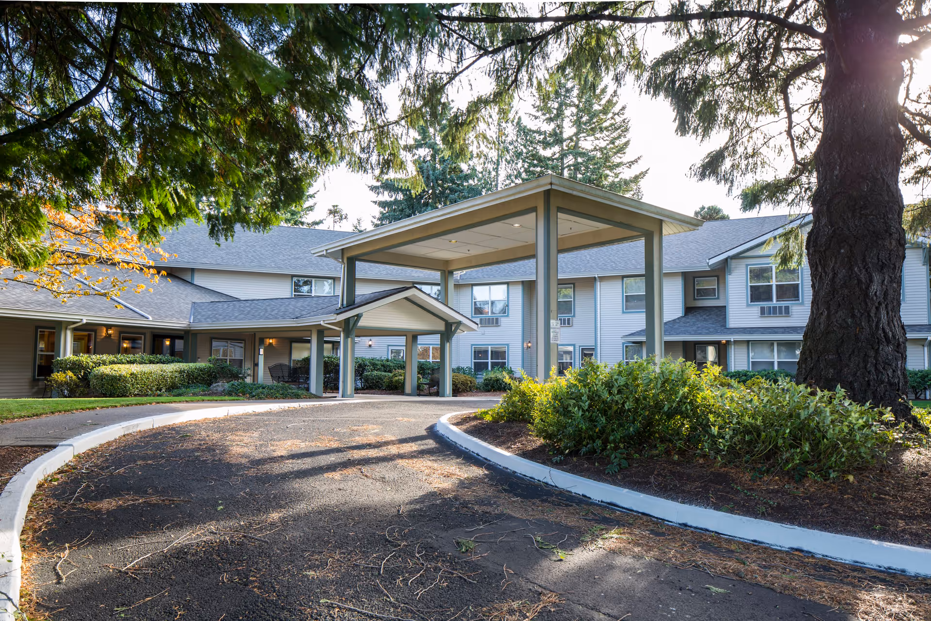 Exterior view of Markam House senior living facility showing a two-story building with a covered entrance driveway surrounded by trees and landscaped bushes.