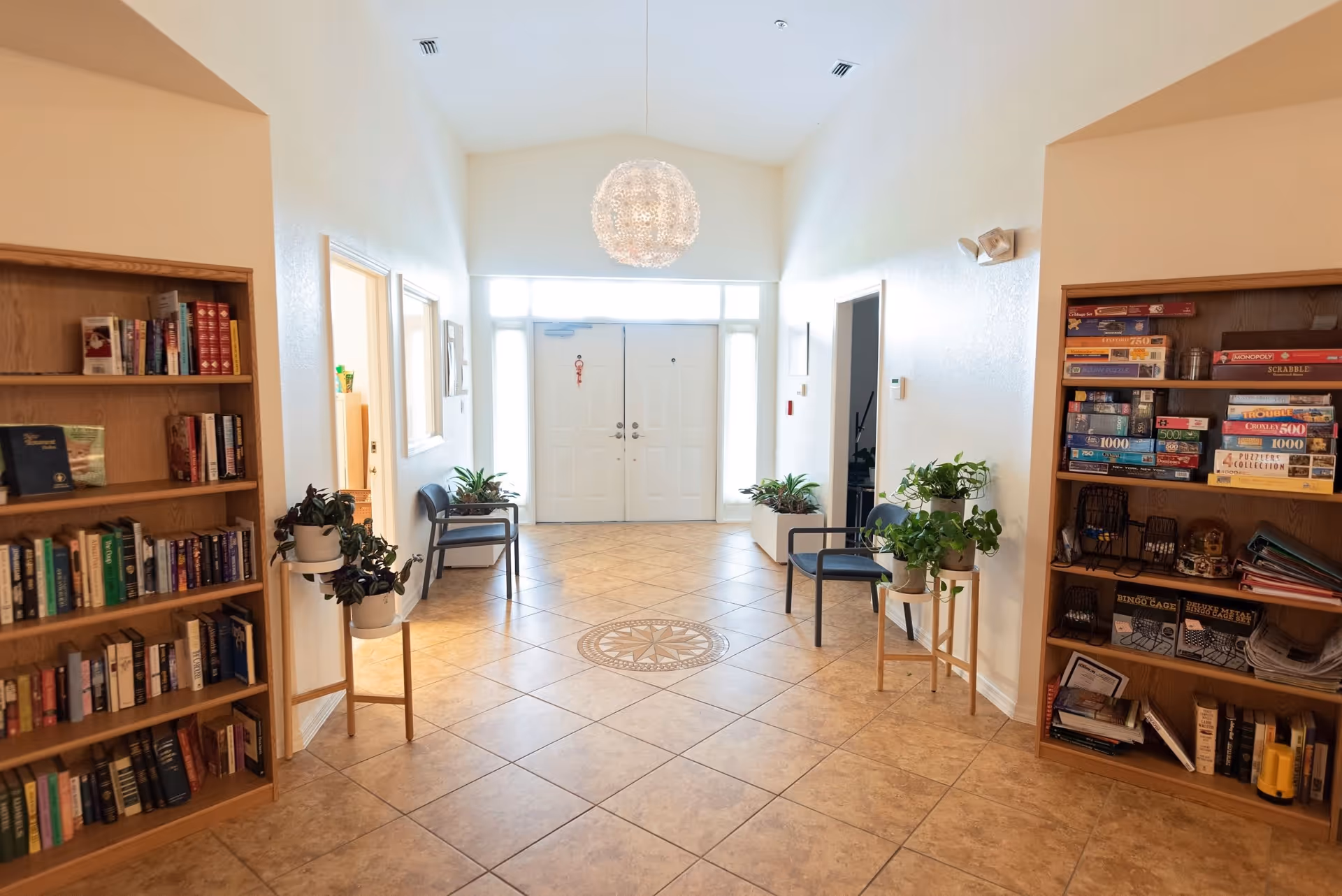 A bright hallway with tiled floor and a decorative circular tile pattern in the center. There are two wooden bookshelves filled with books and board games on either side. Several potted plants are placed on stands and near chairs along the walls. At the end of the hallway, there is a set of double white doors with windows above and on the sides, allowing natural light to enter. A modern spherical chandelier hangs from the ceiling.