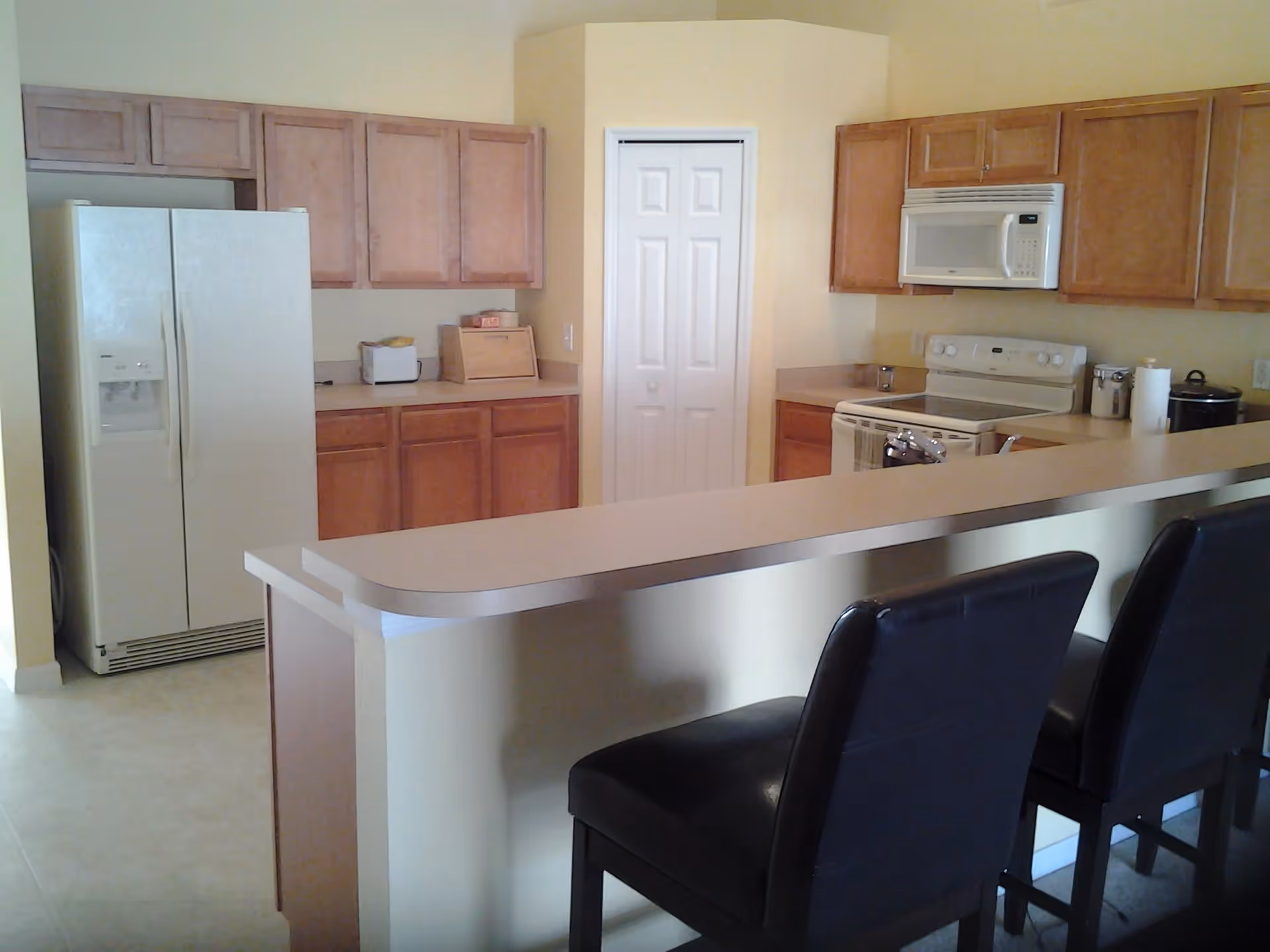 A kitchen area with wooden cabinets, a white refrigerator, a white microwave above a white stove, and a countertop with two black bar stools in front.