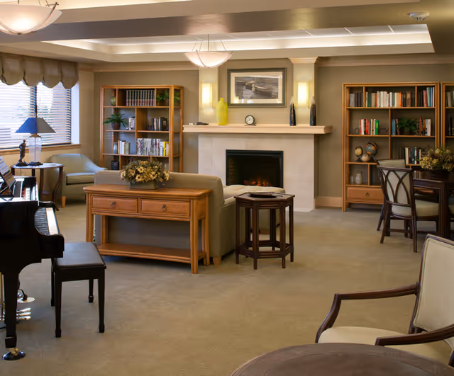 A cozy living room area with a fireplace in the center, flanked by two wooden bookshelves filled with books and decorative items. There is a sofa with a wooden console table behind it, a small side table, and several chairs arranged around the room. A piano with a bench is visible on the left side near a window with blinds and a lamp. The room is warmly lit with ceiling lights and wall sconces.