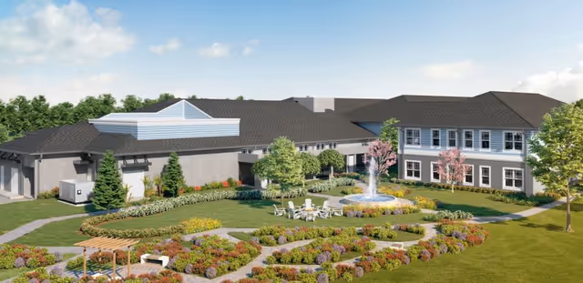 Exterior view of HearthStone at Nona Lakes facility showing a large building with multiple windows surrounded by landscaped gardens, walking paths, trees, and a central water fountain with seating area under a clear sky.