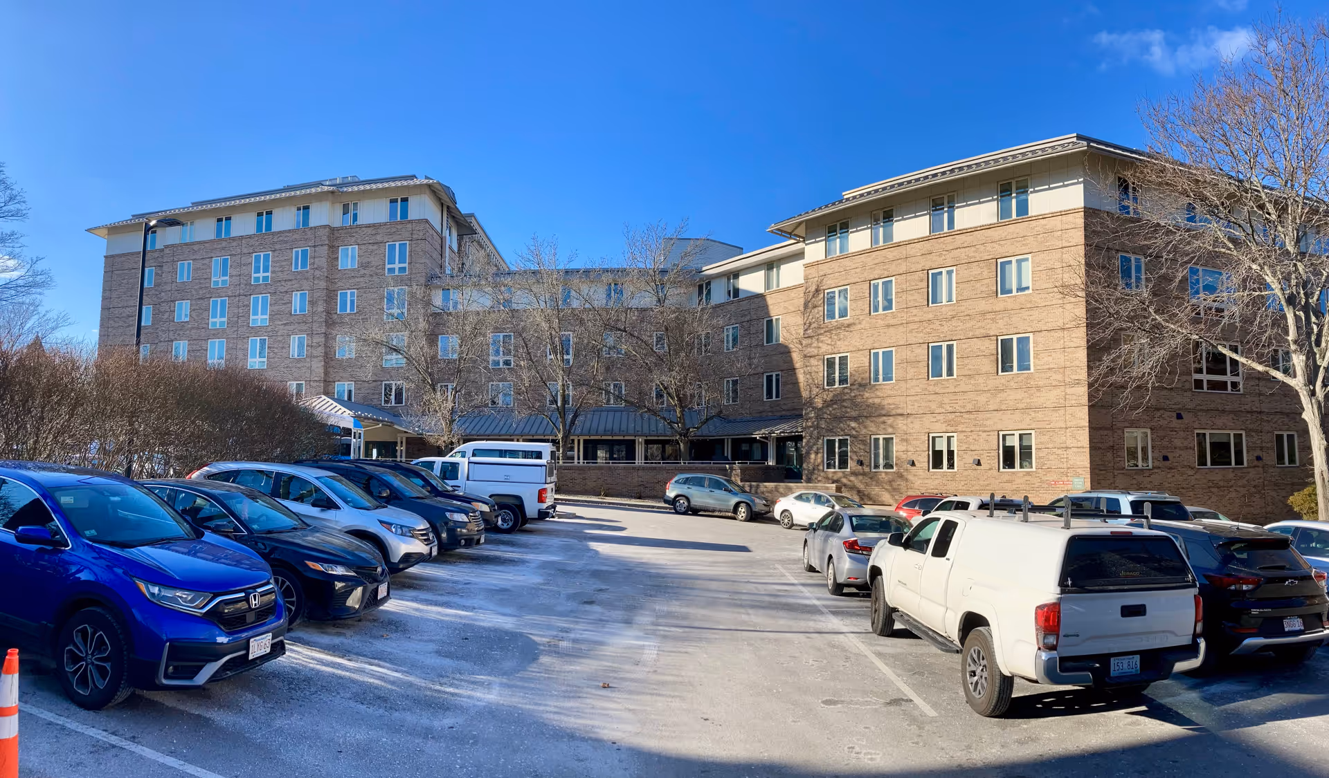 Exterior view of a multi-story brick building with numerous windows under a clear blue sky. In front of the building is a parking lot with several parked cars and a few leafless trees.