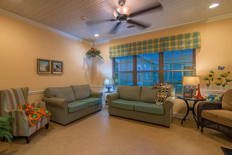 A cozy living room area with two green sofas, a striped armchair with a colorful crocheted blanket, and a wicker chair. The walls are painted light yellow with sunflower and butterfly decals on one side. There are two framed pictures on one wall, a hanging plant in the corner, and a window with a yellow and green checkered valance. Two table lamps sit on side tables beside the sofas, and a ceiling fan with a light fixture is mounted on the wooden paneled ceiling.
