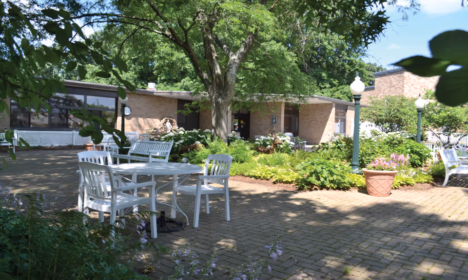 Outdoor patio area at Vincentian Home with white plastic chairs and tables arranged on a brick-paved surface, surrounded by green plants, trees, and flower pots. A single-story brick building is visible in the background under a clear blue sky.