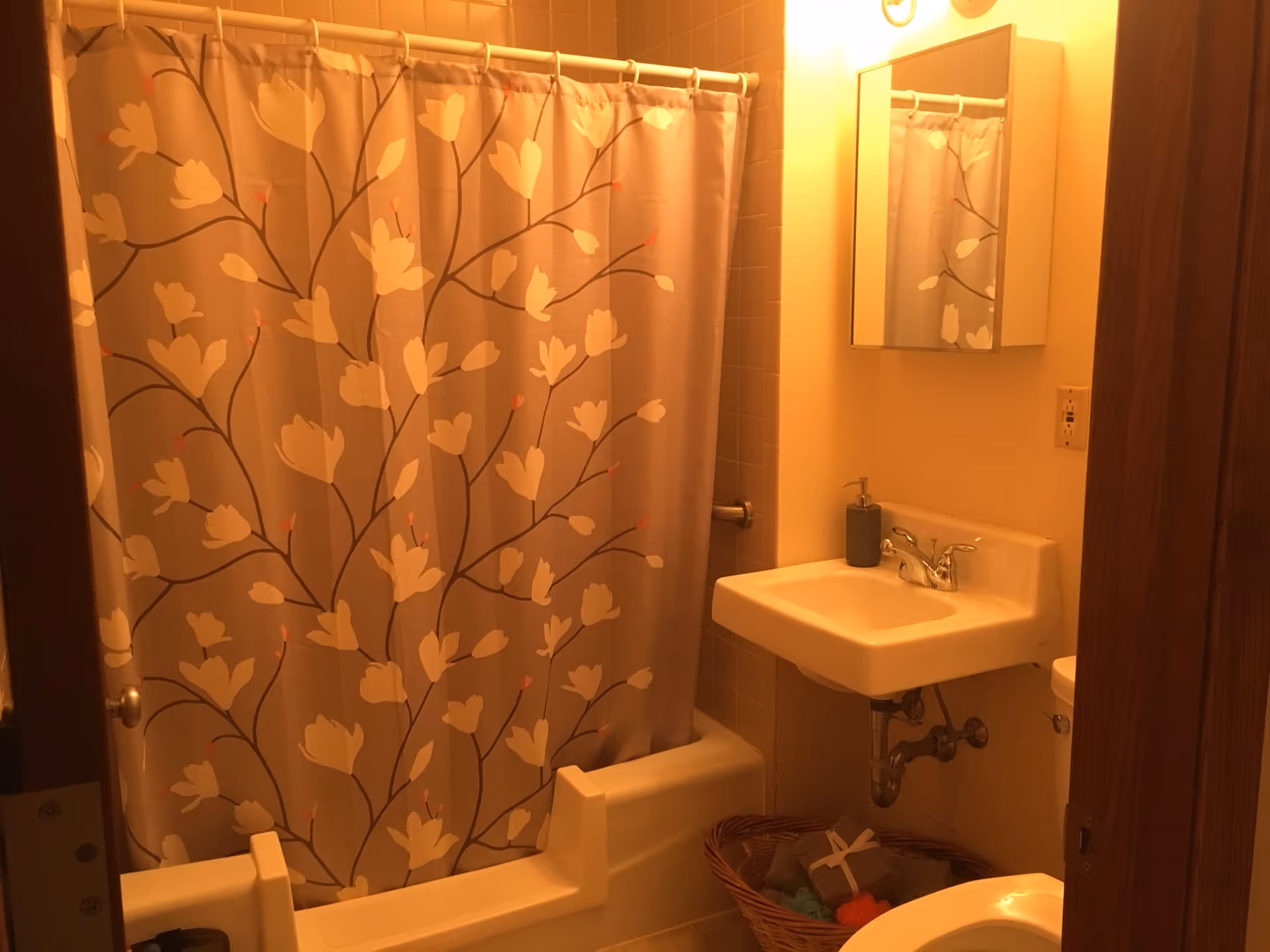 A small bathroom with a floral patterned shower curtain, a white sink with a soap dispenser, a mirror above the sink, and a wicker basket with towels underneath the sink. The lighting is warm and the walls are light-colored.
