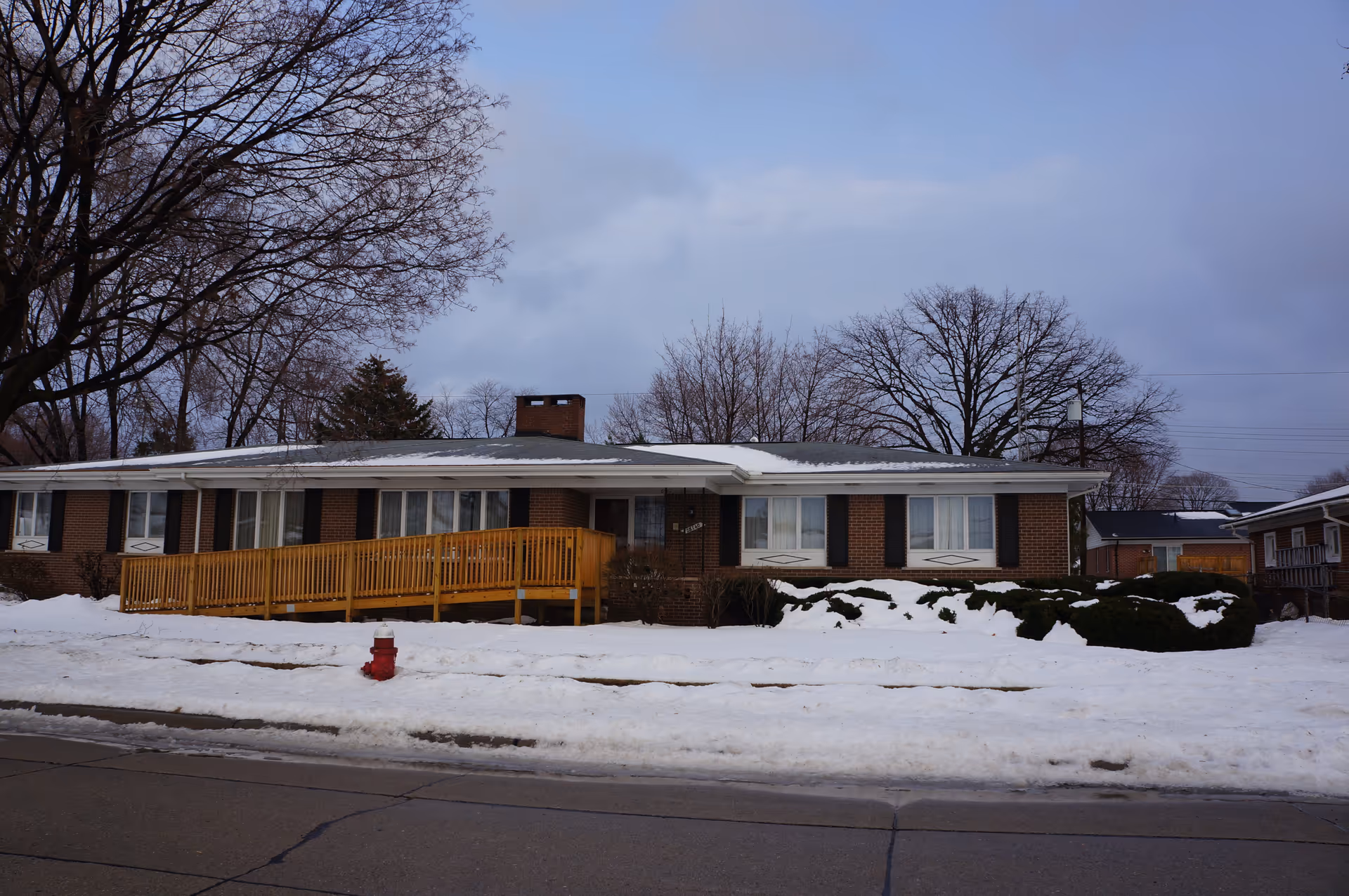Single-story brick building with a wooden wheelchair ramp leading to the entrance, surrounded by snow-covered ground and leafless trees under a cloudy sky.