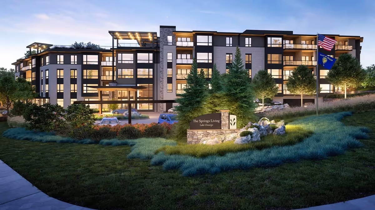 Exterior view of The Springs Living at Lake Oswego, a modern multi-story senior living facility with large windows and balconies, surrounded by landscaped greenery, trees, and a well-lit entrance sign. Two flags, including the American flag and the State of Oregon flag, are visible on flagpoles near the building.
