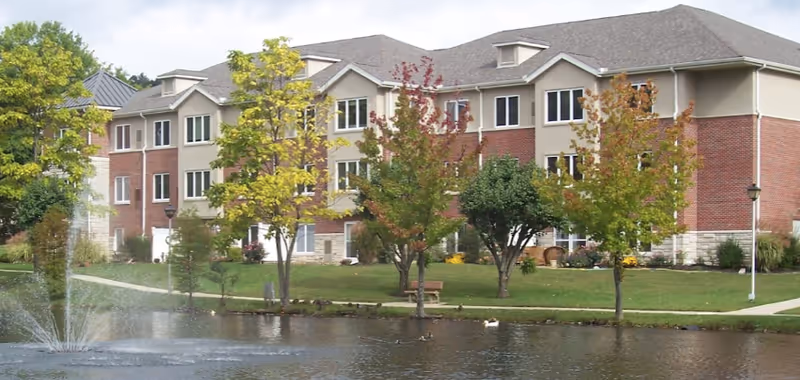 Exterior view of a multi-story retirement center building with brick and beige siding, surrounded by trees with autumn foliage, a pond with a water fountain, and a walking path with benches.