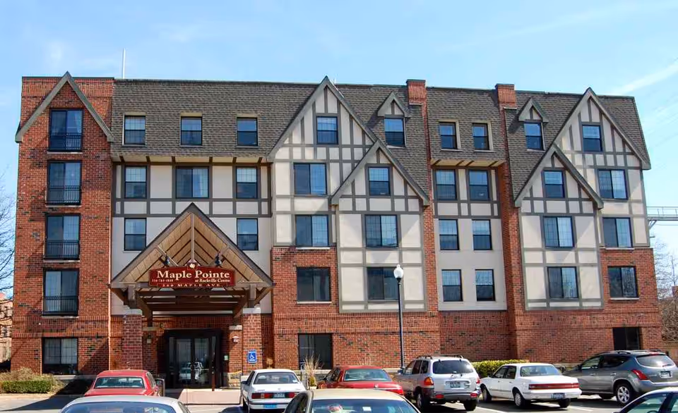 Front exterior view of a multi-story senior living facility named Maple Pointe, featuring a Tudor-style architectural design with brick and timber framing. Several cars are parked in front of the building under a clear blue sky.
