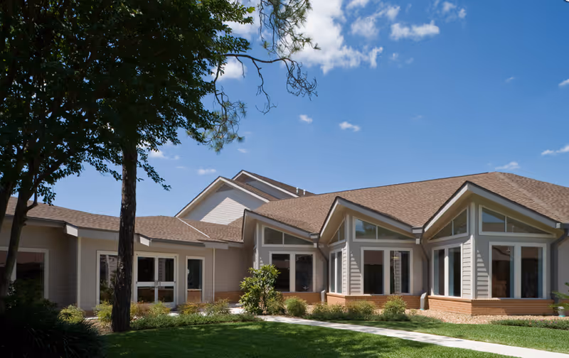 Exterior view of a single-story building with beige siding and brown shingled roof under a clear blue sky. The building is surrounded by green grass, bushes, and trees, with a concrete walkway leading to the entrance.