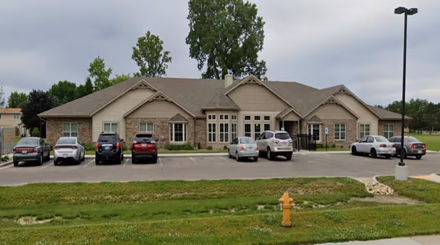 Front exterior view of a single-story assisted living facility building with a beige and brick facade, several windows, and a parking lot with cars parked in front. There is a grassy area with a yellow fire hydrant in the foreground and a tall street lamp on the right side.