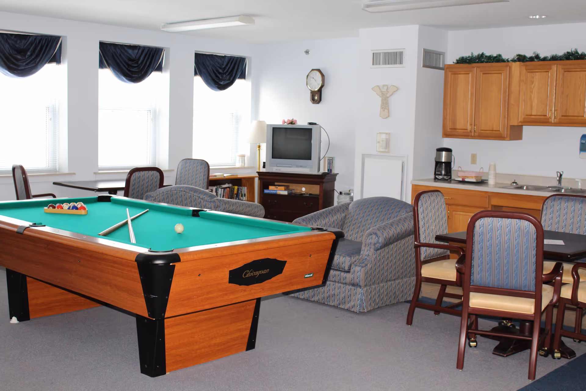 A recreational room in a senior living facility featuring a pool table with cues and balls set up, several upholstered armchairs, a small dining table with chairs, a television on a wooden stand, and a kitchenette area with wooden cabinets, a coffee maker, and a sink. Three windows with dark valances allow natural light into the room.