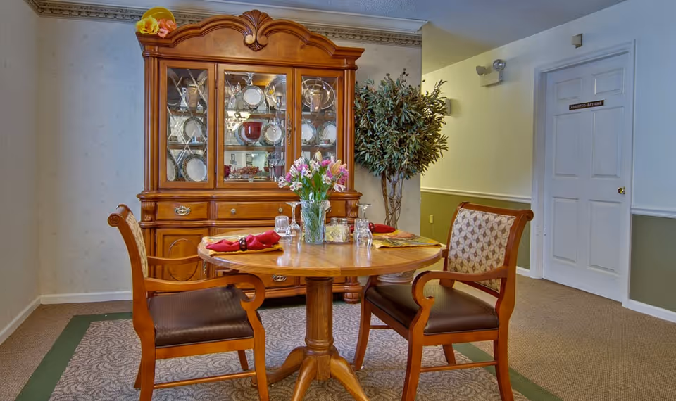 A small dining area with a round wooden table set for two with red napkins and glassware. Behind the table is a wooden china cabinet displaying plates and glassware. A potted plant is placed next to the cabinet, and a door labeled 'Restricted Bathing' is visible on the right side of the image.