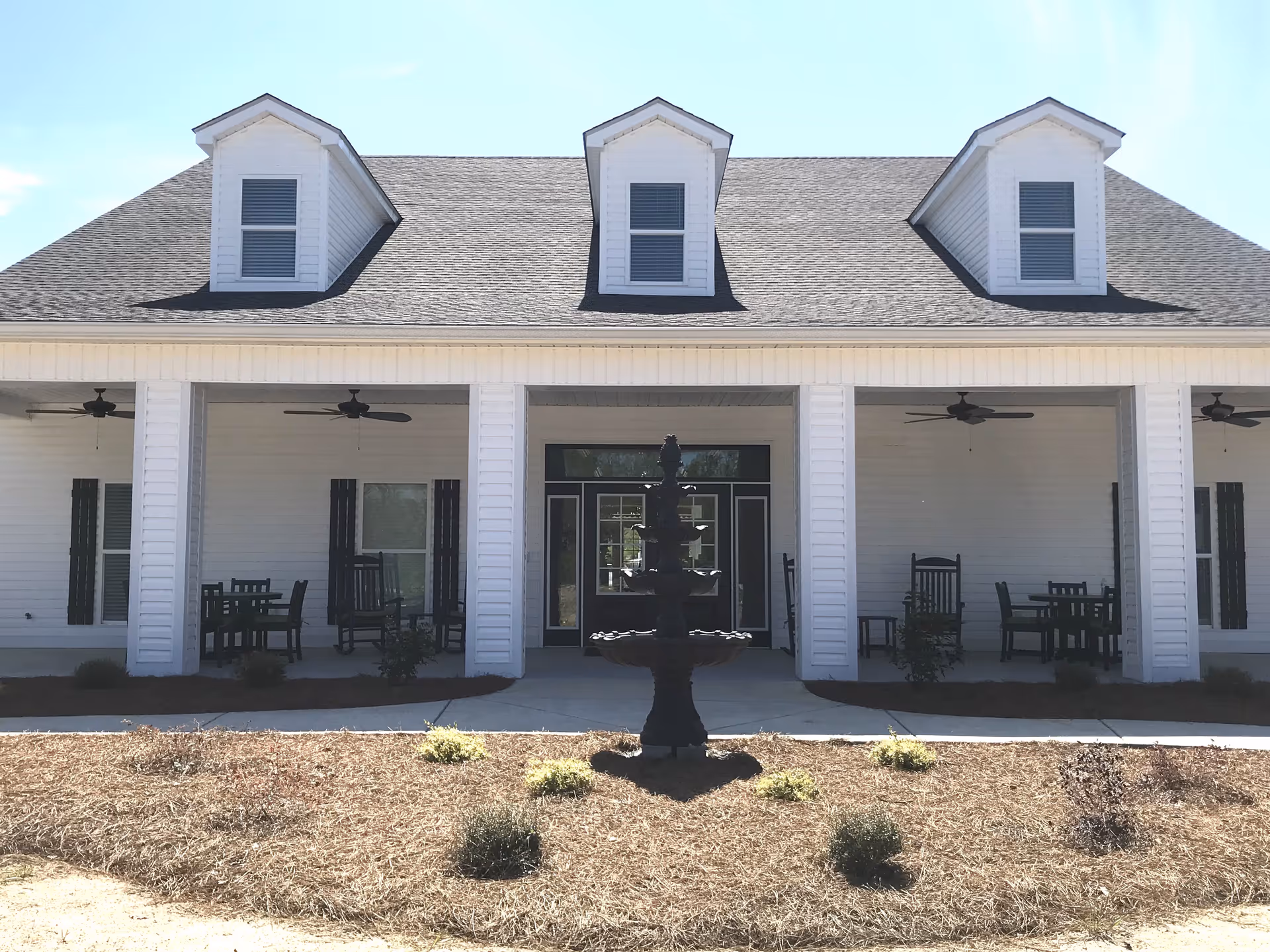 Front exterior view of a white senior living facility building with three dormer windows on the roof, a covered porch with ceiling fans, rocking chairs, and tables. In front of the building is a black multi-tiered fountain surrounded by small plants and mulch.