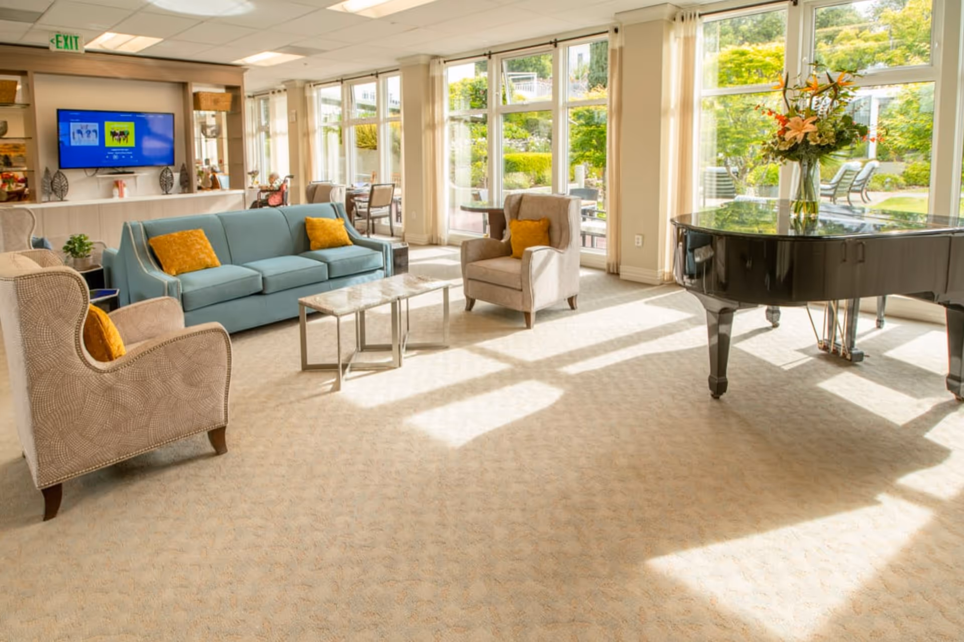 Bright senior living common area with large windows letting in natural light, featuring a blue sofa with yellow pillows, two beige armchairs with yellow pillows, a black grand piano with a vase of flowers on top, and a TV mounted on the wall. Outside the windows, greenery and patio furniture are visible.