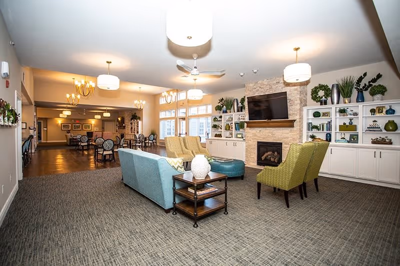 A spacious senior living common area with a seating arrangement including a blue sofa, green armchairs, and a round ottoman. The room features a stone fireplace with a mounted TV above it, built-in white shelves decorated with plants and ornaments, and large windows letting in natural light. In the background, there is a dining area with multiple tables and chairs under modern chandeliers.