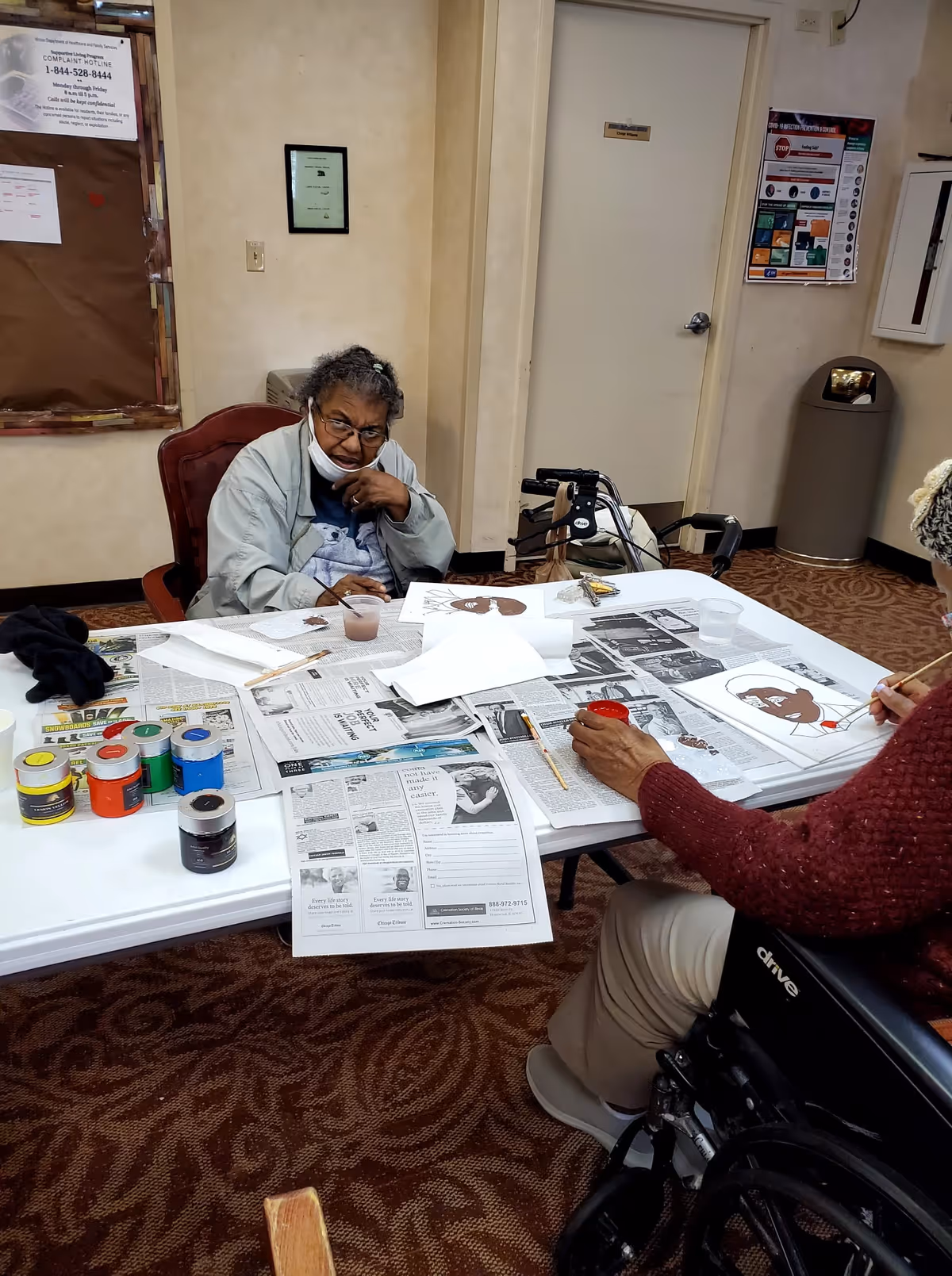 Two elderly individuals seated at a table covered with newspapers and art supplies, including paint jars and brushes, engaged in painting activities inside a room with beige walls and carpeted floor. One person is wearing a light jacket and glasses, while the other is in a wheelchair wearing a red sweater.