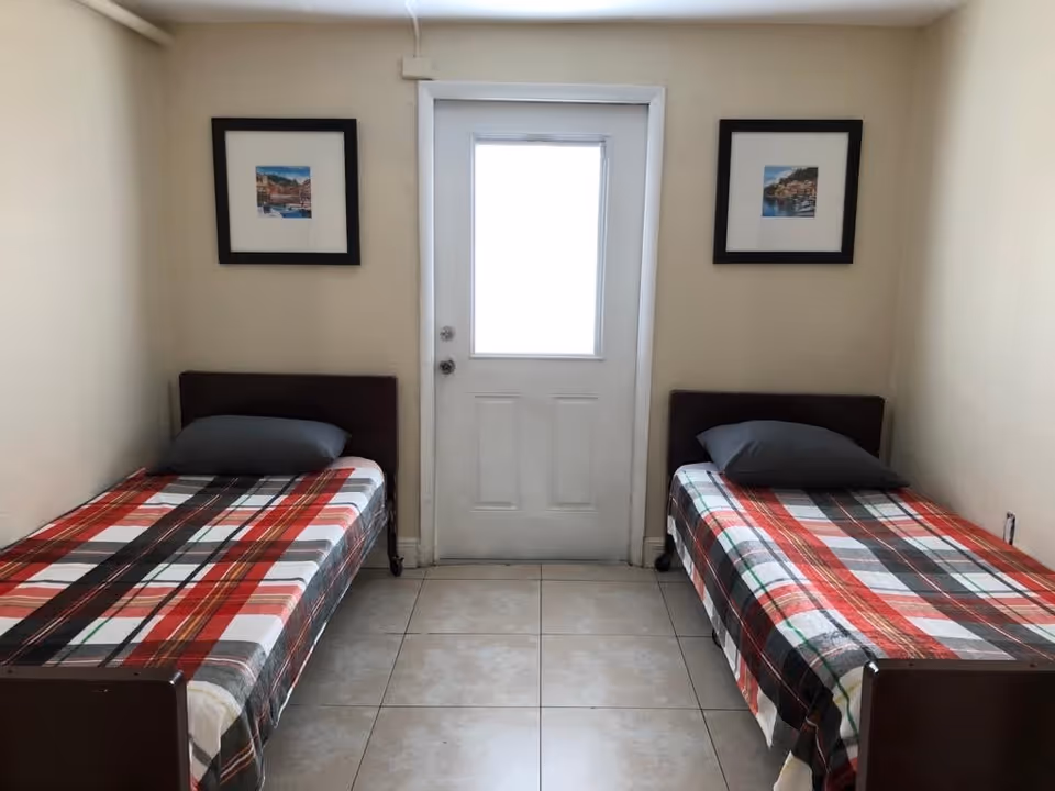 A small room with two single beds placed on opposite sides, each with a plaid blanket and a gray pillow. A white door with a window is centered on the back wall between the beds. Two framed pictures hang on the wall above each bed. The floor is tiled and the walls are painted beige.