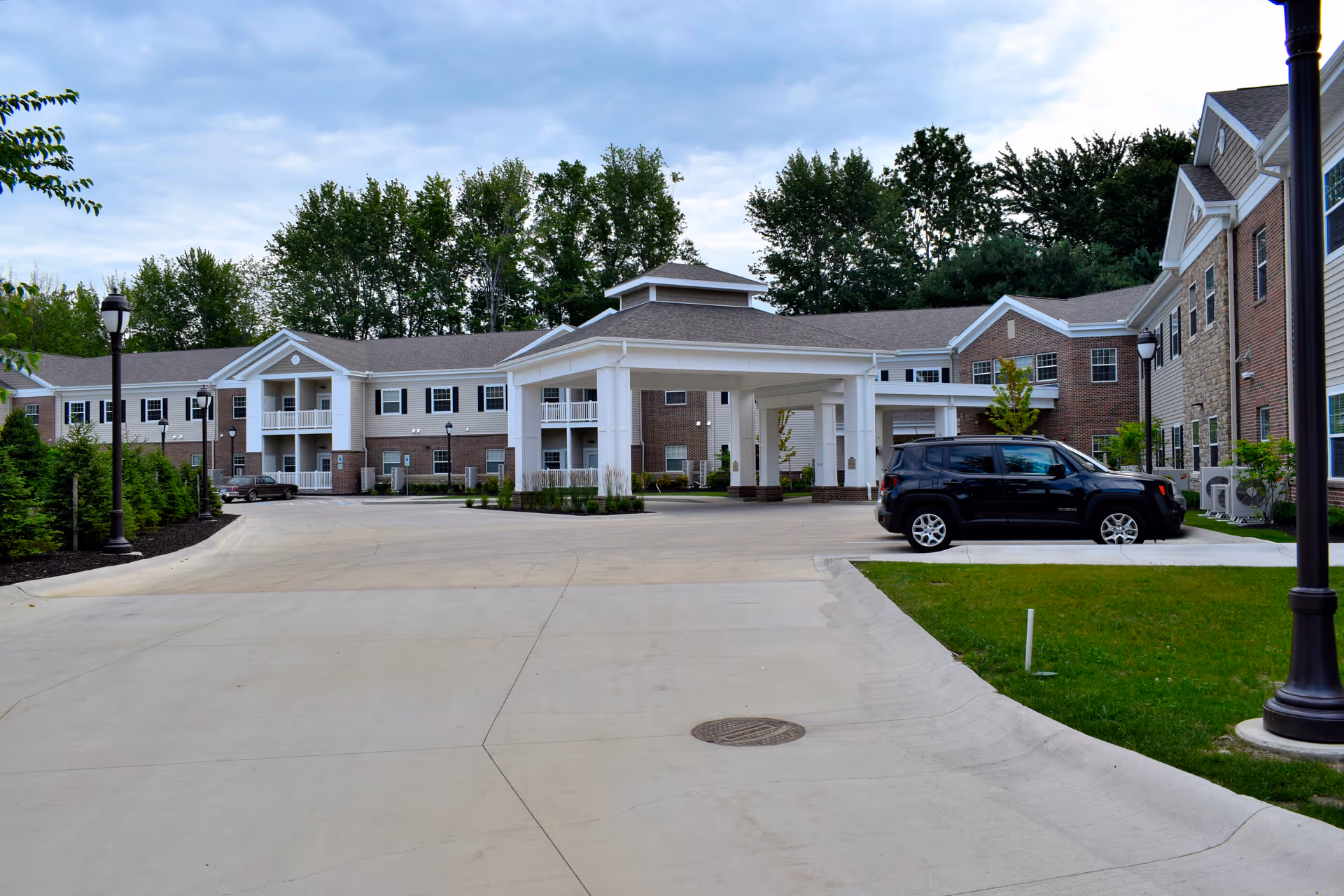 Exterior view of Danbury Mentor senior living facility showing a large driveway with a covered entrance, surrounded by a two-story building with brick and siding facade, several windows, and balconies. There are a few parked cars, street lamps, green grass, and trees in the background under a cloudy sky.