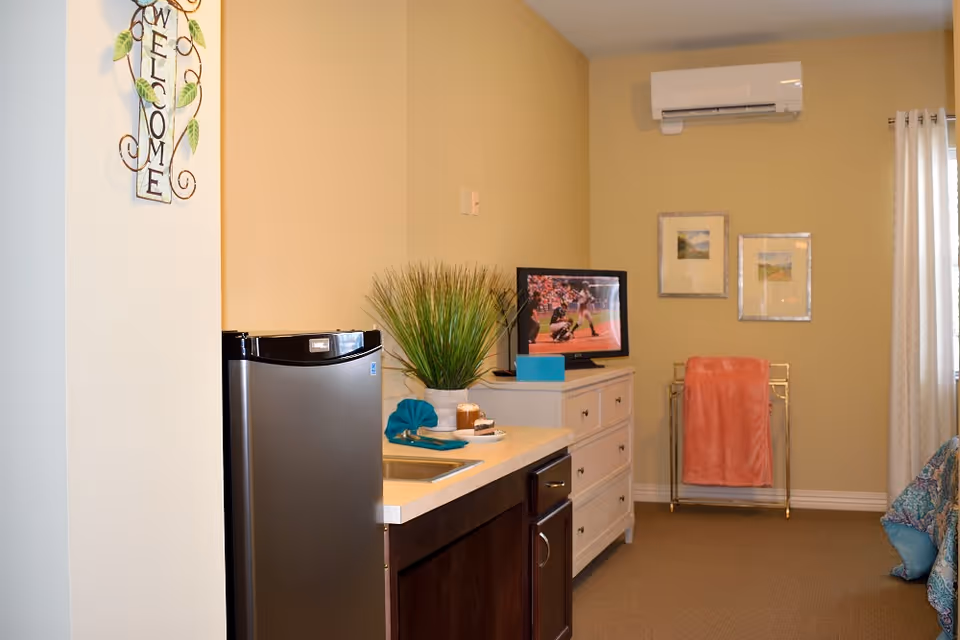 Interior view of a cozy room with a small kitchenette featuring a mini fridge, sink, and countertop. A white dresser with a TV on top is against the wall, displaying a sports game. Above the dresser are two framed pictures. A coral-colored towel hangs on a gold towel rack near a window with white curtains. A decorative welcome sign is mounted on the wall near the kitchenette.