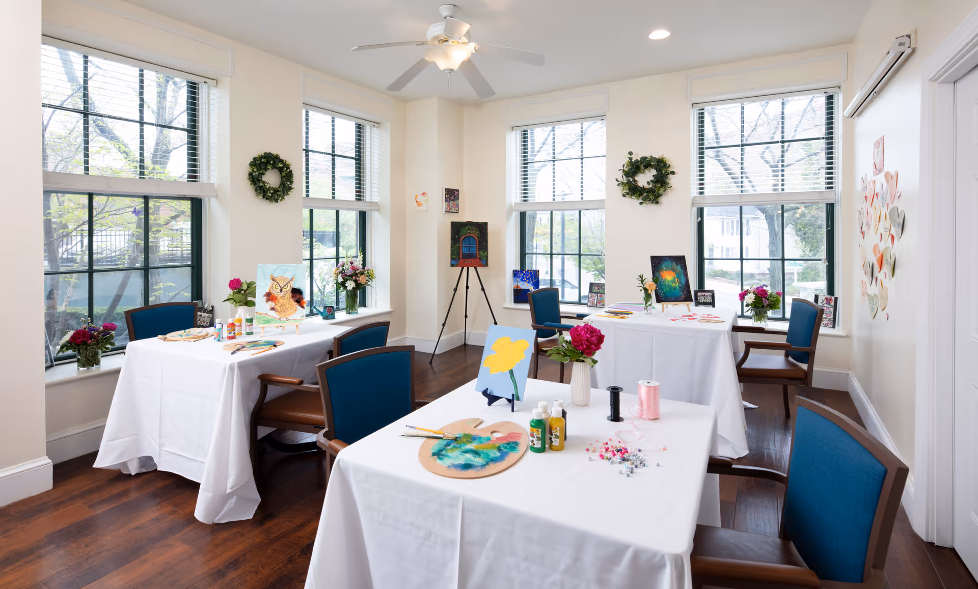 A bright and airy activity room with large windows letting in natural light. The room has three tables covered with white tablecloths, each set up for painting and crafts with paint bottles, brushes, palettes, and small art pieces. Blue cushioned chairs surround the tables. The walls are decorated with wreaths and small artworks, and there are flowers in vases on the tables and window sills.