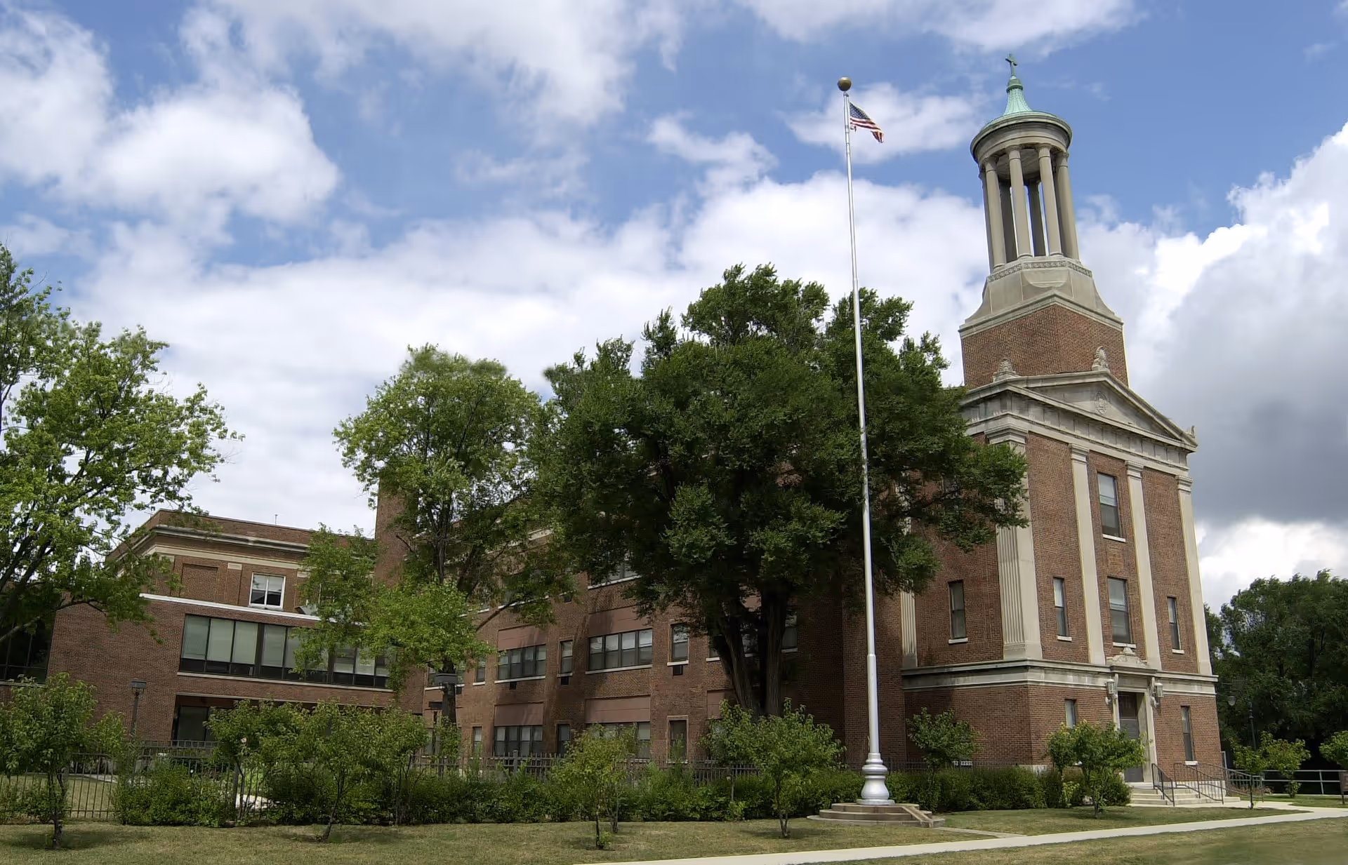 Exterior view of a large brick building with a tall tower and an American flag on a flagpole in front. The building is surrounded by green trees and bushes under a partly cloudy sky.
