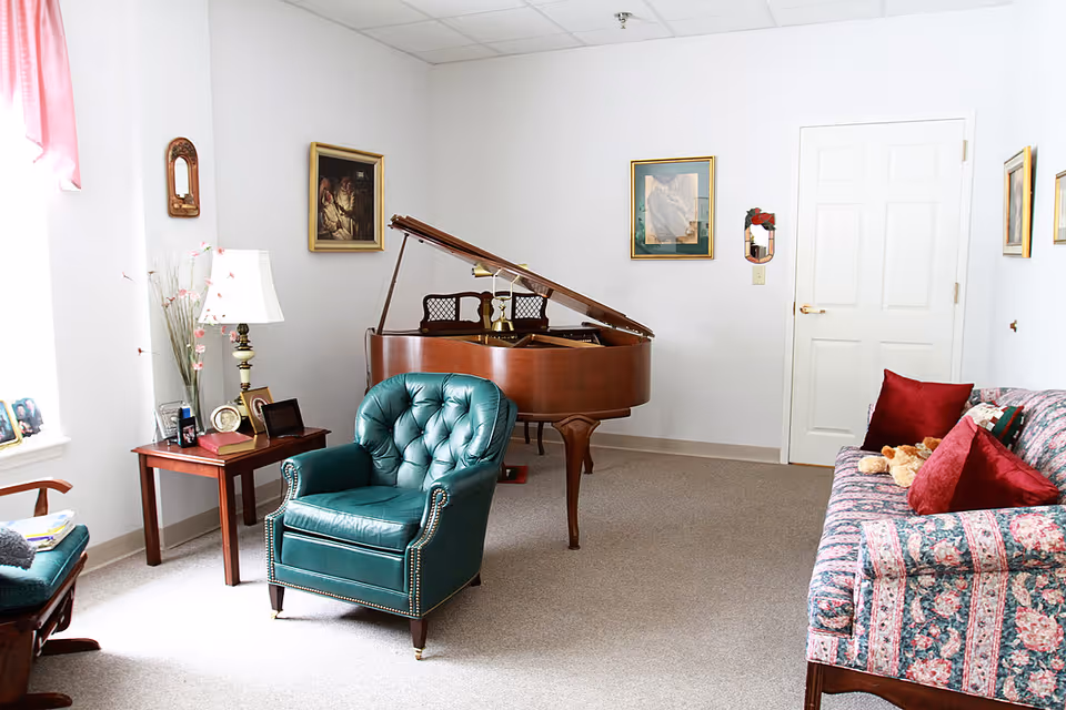 A cozy living room with a green leather armchair, a floral patterned sofa with red pillows, a wooden grand piano, a wooden side table with a lamp and framed photos, and several framed paintings on the white walls.