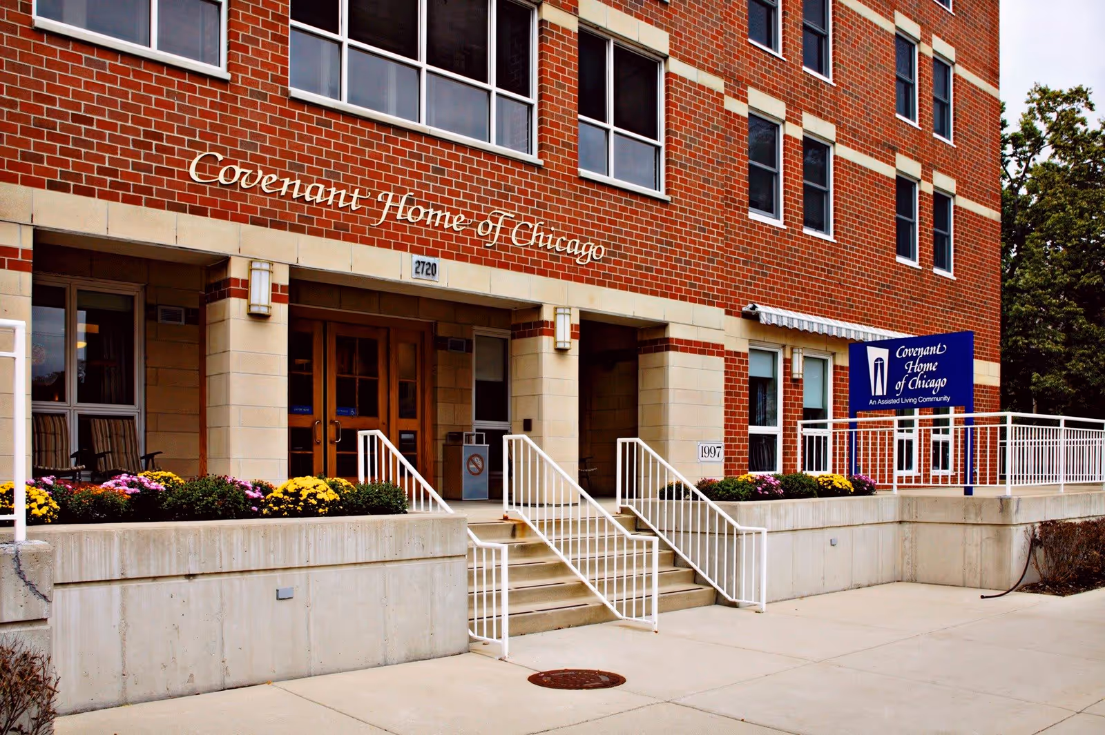 Exterior front entrance of Covenant Home of Chicago, a brick building with large windows, stairs with white railings leading to double doors, flower beds with colorful flowers, and a blue sign indicating it is an assisted living community.