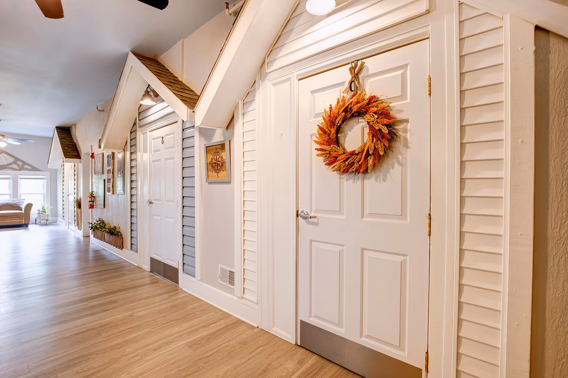 A bright hallway in a senior living facility with doors designed to look like small houses, each with a peaked roof and siding. One door is decorated with an autumn-themed wreath. The hallway has light wood flooring, a ceiling fan, and a seating area with a couch and windows at the far end.