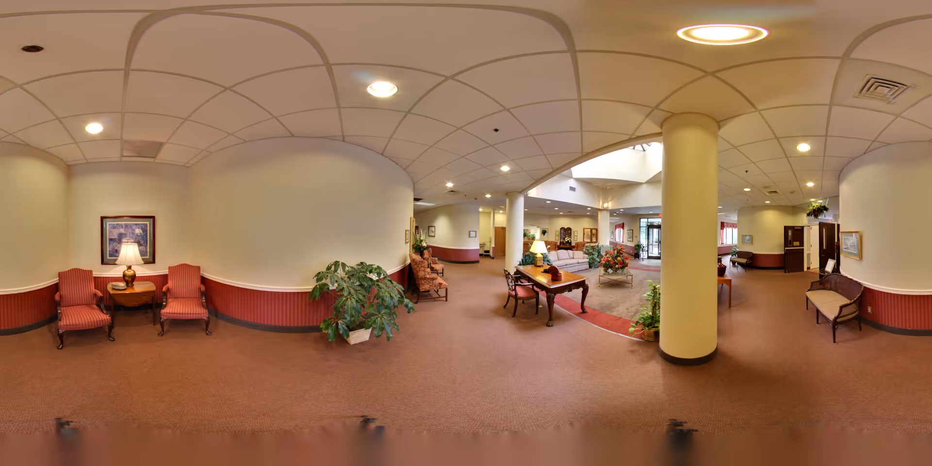A spacious senior living facility lobby with beige walls and carpeted floors. The area features several seating arrangements including red upholstered chairs, wooden benches, and a sofa. There are decorative plants, a table with a lamp, framed artwork on the walls, and a large column in the center. The entrance doors are visible in the background with natural light coming through.