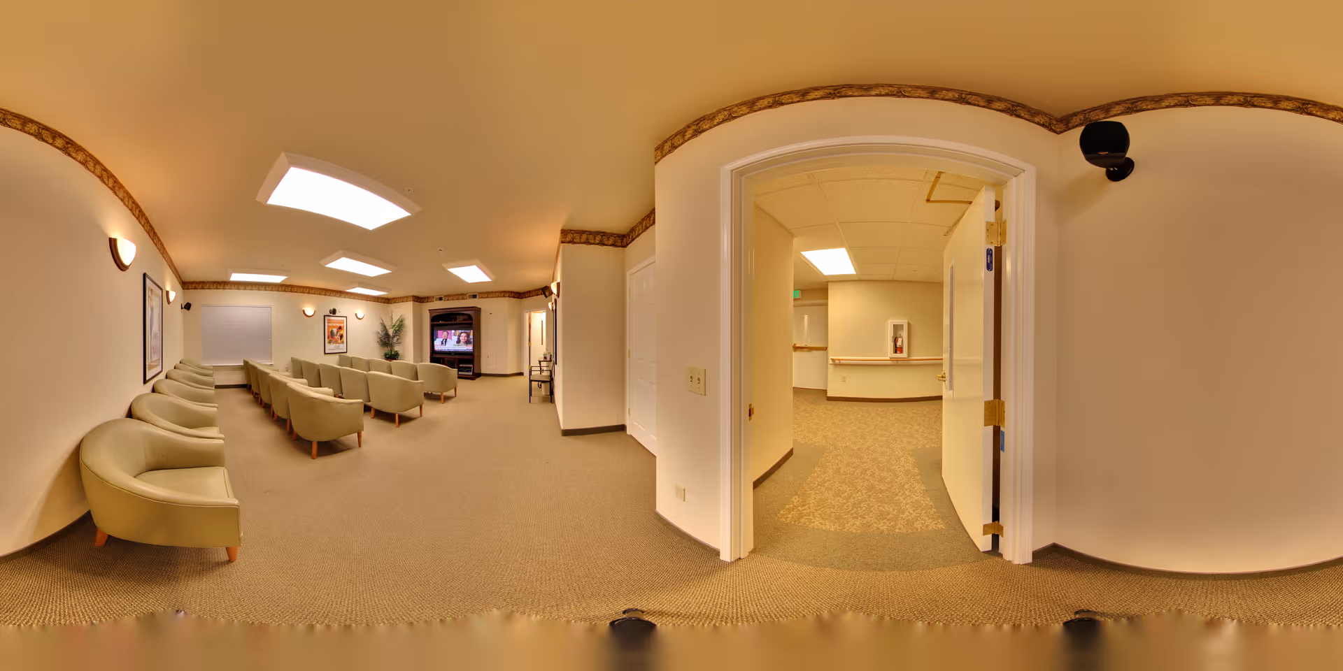 A spacious, well-lit common area with rows of beige armchairs facing a large television mounted on the far wall. The room has beige walls, carpeted floors, and decorative crown molding near the ceiling. To the right, an open doorway leads to a hallway with similar carpeting and lighting.