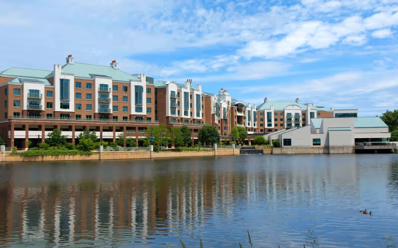 A large multi-story senior living facility building with green roofs and brick exterior, situated beside a calm body of water reflecting the building and blue sky with scattered clouds.