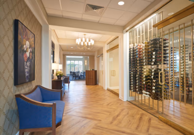 Interior hallway of a senior living facility with wooden flooring and a blue cushioned bench on the left. A framed floral painting hangs above the bench. On the right, there is a glass-enclosed wine storage area with multiple bottles of wine displayed. At the end of the hallway, a dining area with tables and chairs is visible under a chandelier.