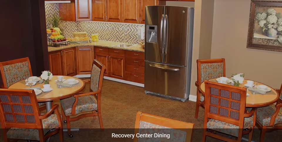 Dining area in a recovery center featuring round wooden tables set with plates, cups, and small flower arrangements. The background shows a kitchen area with wooden cabinets, a mosaic tile backsplash, a double sink, and a stainless steel refrigerator. A framed floral painting hangs on the wall.