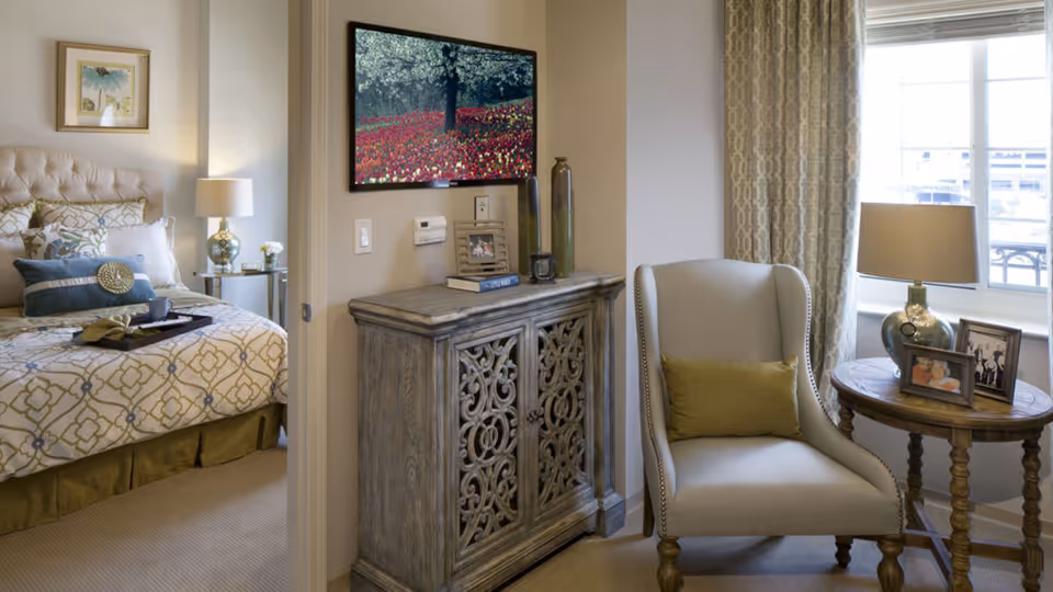 A cozy senior living bedroom with a patterned bedspread, decorative pillows, and a bedside table with a lamp. Adjacent to the bedroom is a small sitting area featuring a cushioned armchair with a green pillow, a round wooden side table with a lamp and framed photos, and a decorative cabinet with intricate carved doors beneath a wall-mounted painting of a tree and red flowers.