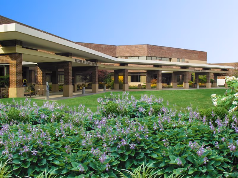 Exterior view of Wexner Heritage Village building with a covered entrance supported by brick columns. In the foreground, there is a well-maintained garden with green plants and purple flowers under a clear blue sky.