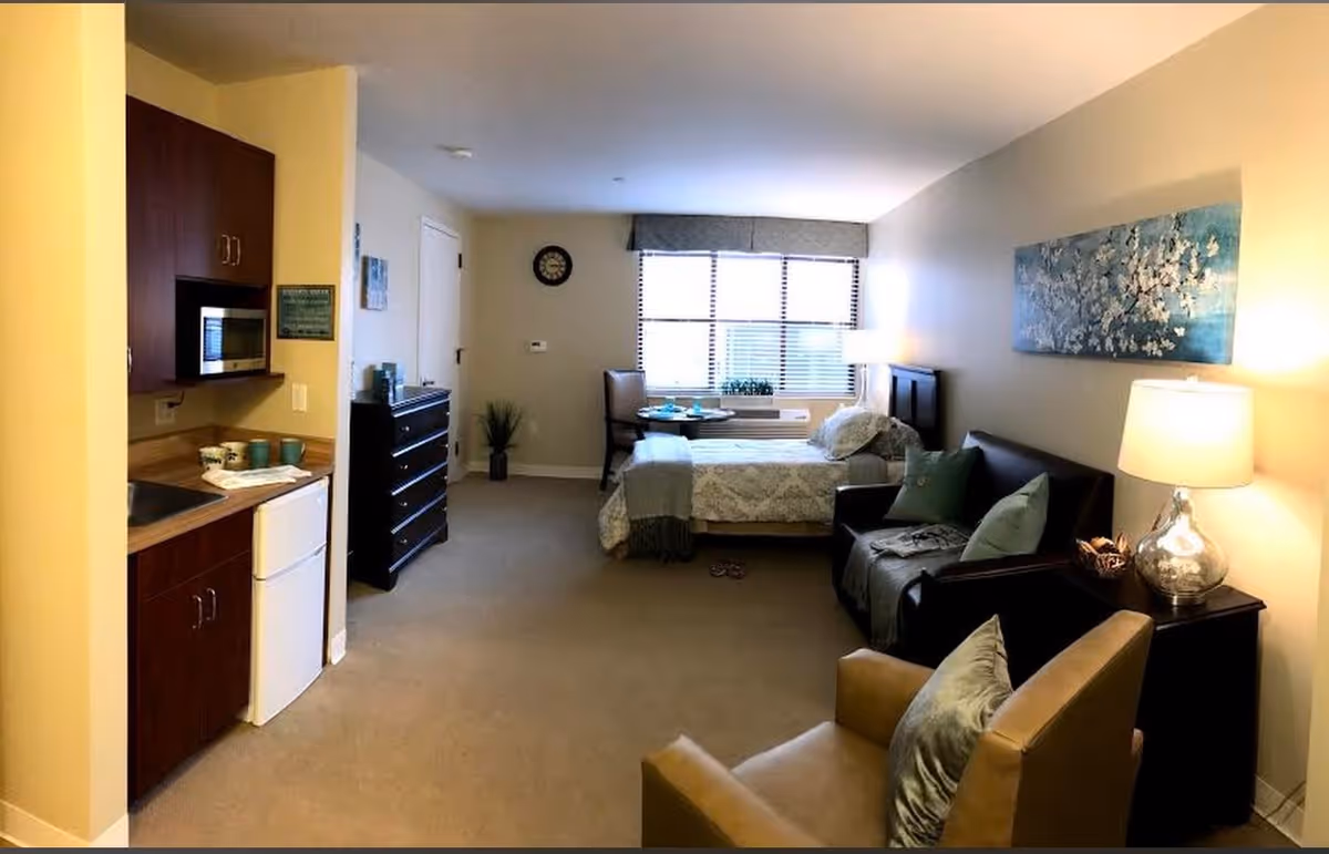 A cozy assisted living studio apartment featuring a small kitchenette with microwave, mini fridge, and cabinets on the left. The room includes a bed with patterned bedding near a window with blinds, a small dining table with two chairs, a dark dresser, a black loveseat with pillows, a beige armchair, a side table with a lamp, and a large floral painting on the wall.