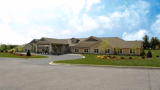 Wide exterior view of a single-story senior living facility building with a covered entrance, surrounded by a well-maintained lawn, small trees, and shrubs under a partly cloudy sky.