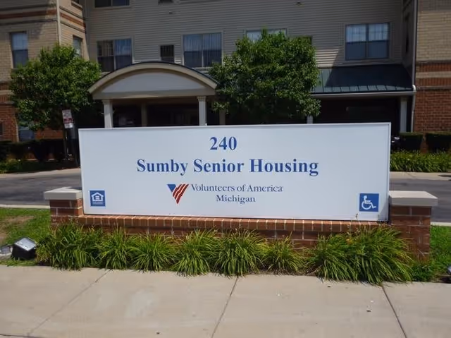 A large white sign in front of a building reads '240 Sumby Senior Housing Volunteers of America Michigan' with logos for equal housing and accessibility. The sign is mounted on a low brick wall with green plants in front and the building entrance visible in the background.