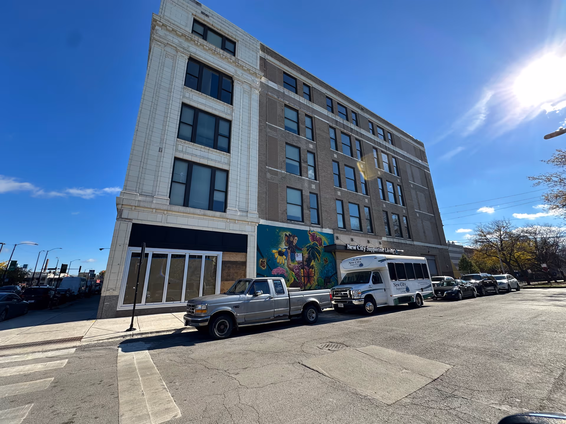 Exterior view of a multi-story building under bright sunlight with a clear blue sky. The building has a mural on one side and a sign that reads 'New City Supportive Living'. Several vehicles, including a pickup truck and a shuttle bus with the same facility name, are parked along the street in front of the building.
