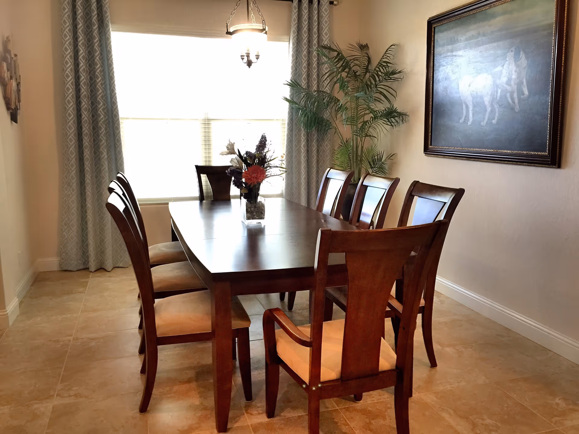 A dining room with a wooden table and eight matching chairs, a vase with flowers on the table, a large window with patterned curtains, a potted plant in the corner, and a framed painting of two white dogs on the wall.